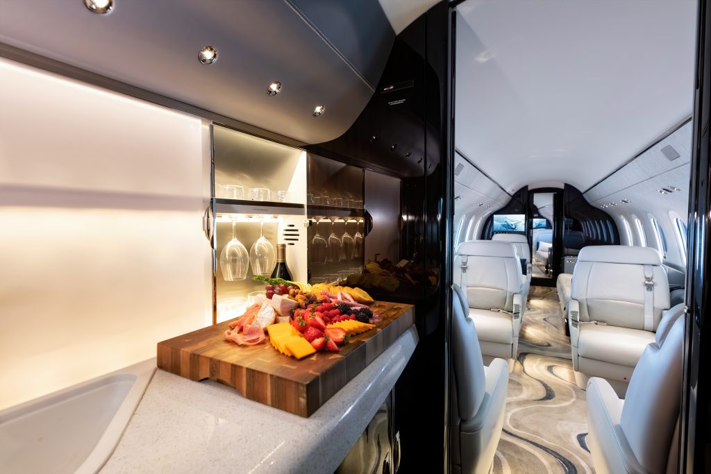 The inside of a private jet with white seats and a bar with wine glasses and a cheese platter with cheese fruit and meat