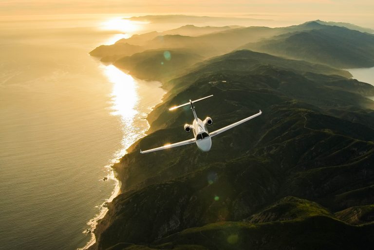 a flying white private jet over sea and mountainous coastline during sunset