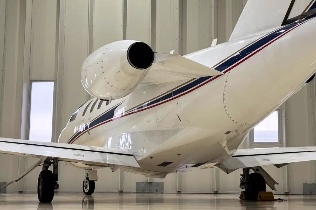 A white private jet with red and blue stripes is parked inside a hangar. The rear of the plane is visible, showcasing its engine and wing.