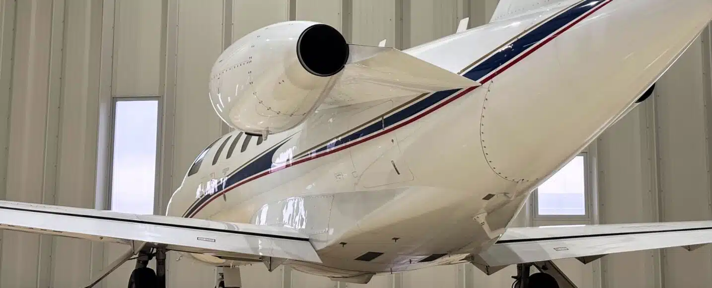 A white private jet with red and blue stripes is parked inside a hangar. The rear of the plane is visible, showcasing its engine and wing.