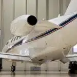 A white private jet with red and blue stripes is parked inside a hangar. The rear of the plane is visible, showcasing its engine and wing.