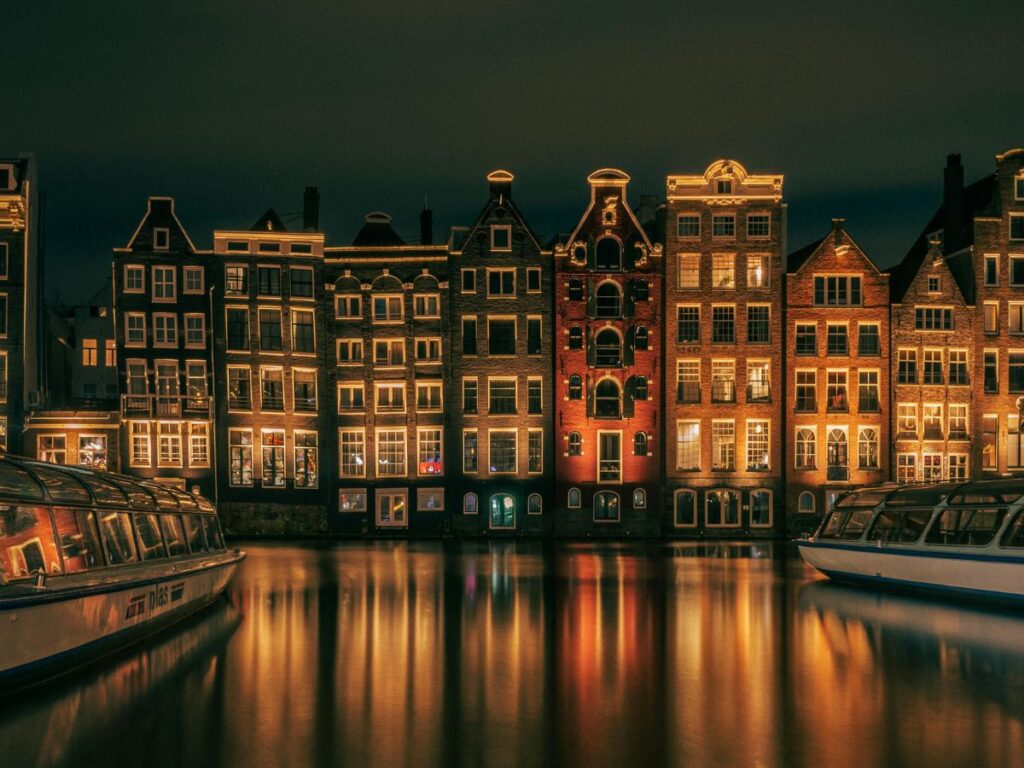 A picturesque view of Amsterdam at night. Several historic buildings with warm, glowing windows line a canal, reflecting their lights on the water. Two boats are moored on the canal, adding to the tranquil scene.