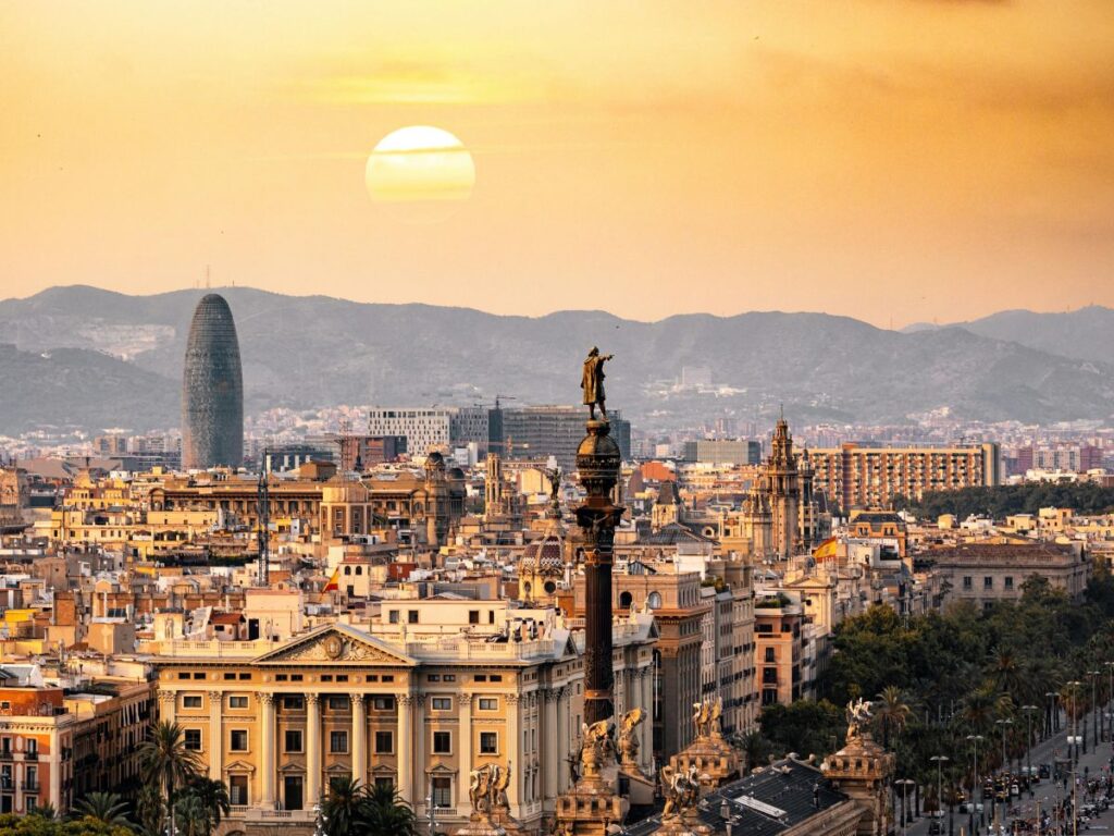 A breathtaking panorama of Barcelona at sunset. The sun sets behind the city, casting a warm glow on the buildings and creating a dramatic silhouette against the sky. The iconic Columbus Monument stands tall in the foreground, overlooking the city. In the distance, the Torre Glòries (Agbar Tower) rises majestically.