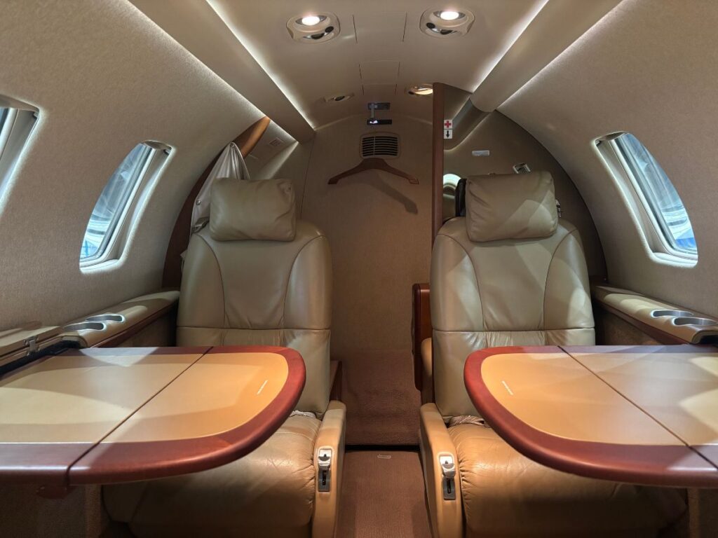 A cozy and well-lit cabin of a private jet. Two plush chairs face each other, each with a small table and drink holder. The interior is beige and brown, creating a warm and inviting atmosphere.