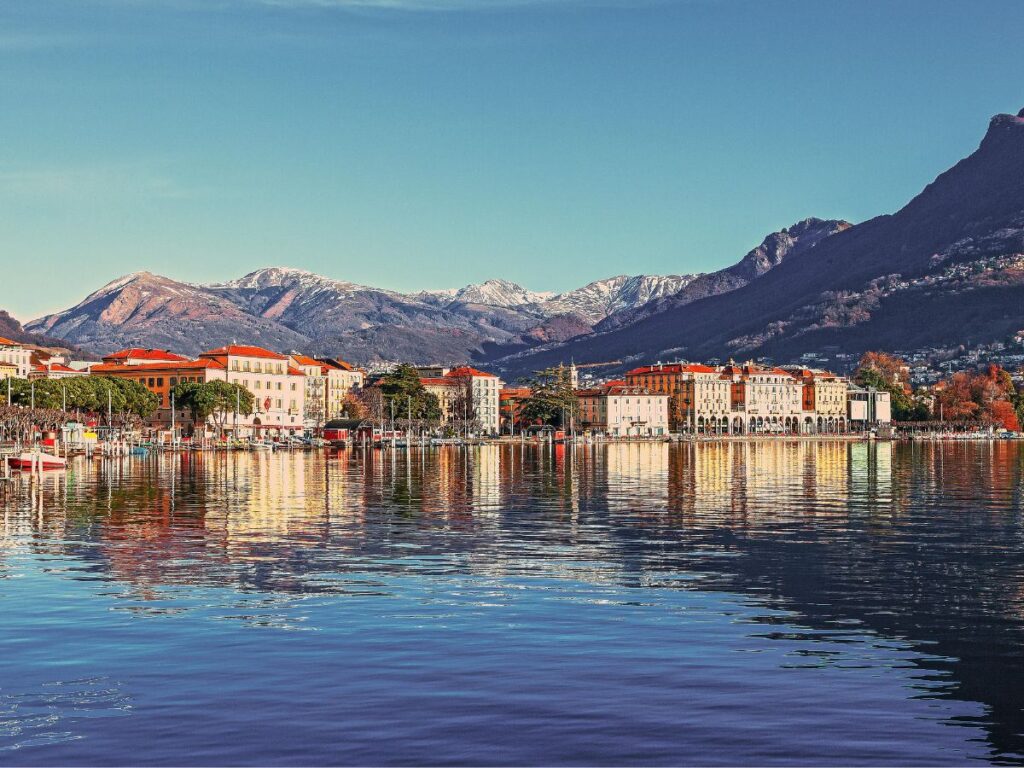 A scenic view of Geneva, Switzerland. The city skyline, with its colorful buildings and architecture, is reflected in the calm water of Lake Geneva. Snow-capped mountains rise majestically in the background, creating a picturesque winter scene.