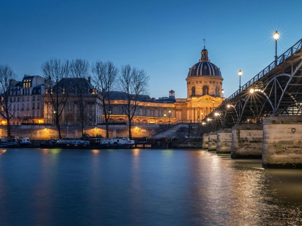 A picturesque view of Paris at dusk. The iconic dome of the Institut de France is illuminated, casting a warm glow on the surrounding buildings and the Seine River. The Pont des Arts bridge, adorned with lampposts, adds a touch of elegance to the scene.