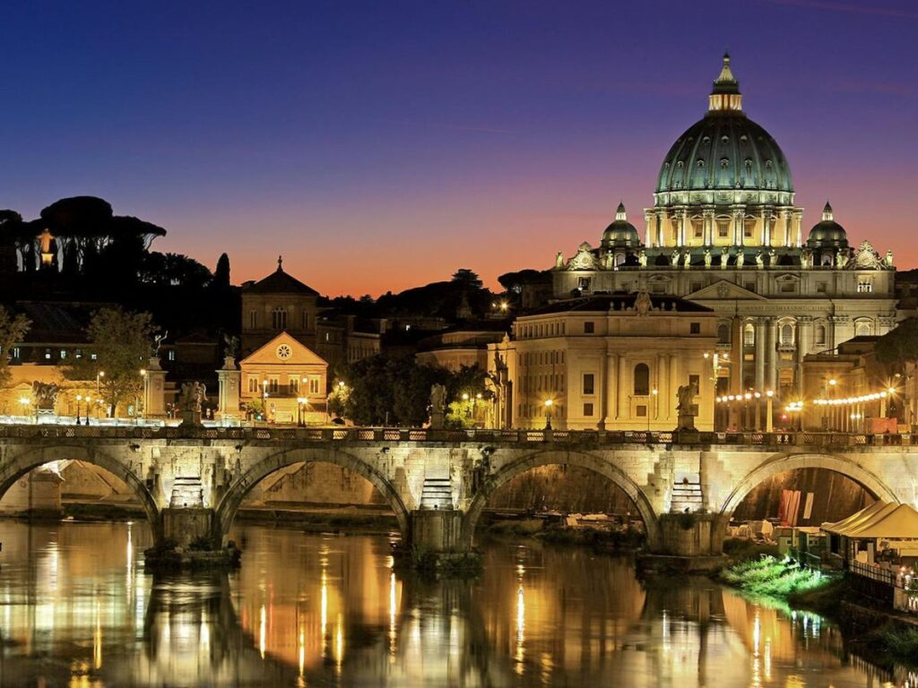 A picturesque view of Rome at dusk. The iconic dome of St. Peter's Basilica and its surrounding buildings are illuminated, casting reflections on the water below. The Ponte Sant Angelo bridge, adorned with statues, adds a touch of grandeur to the scene.