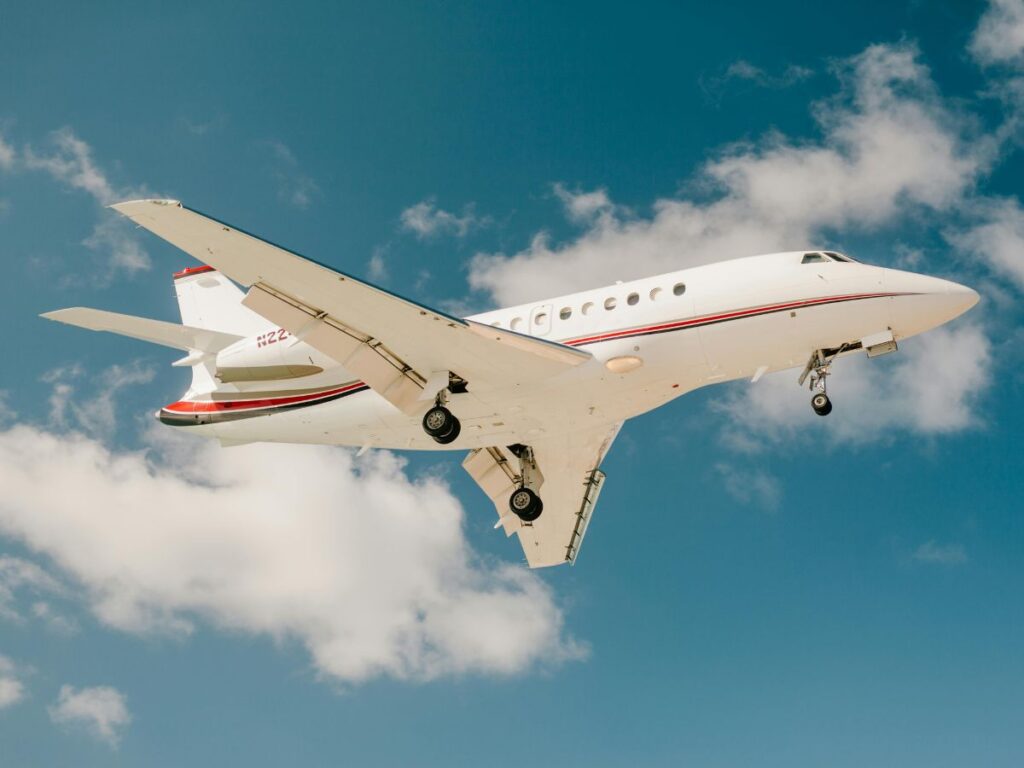 A white private jet with red and blue stripes flies against a clear blue sky with fluffy white clouds. The landing gear is down for landing at the airport.