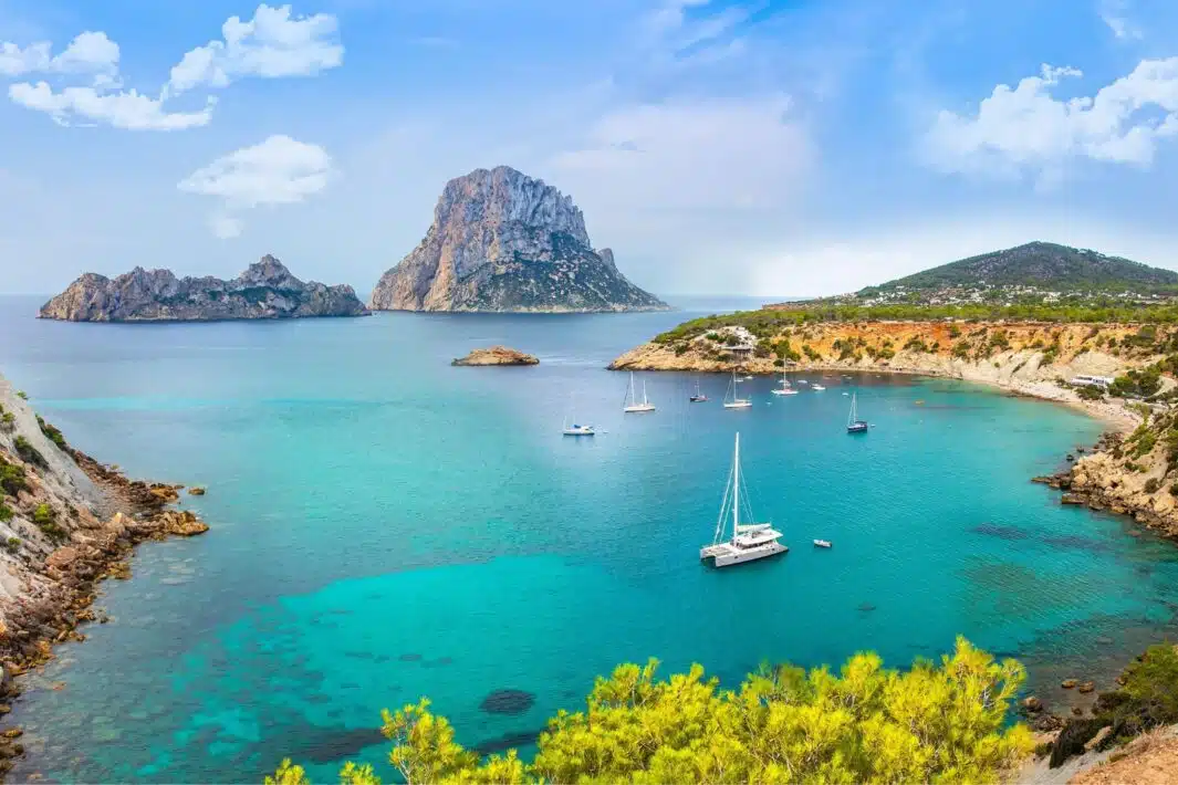 A panoramic view of a picturesque bay in Ibiza, Spain. The clear turquoise water is dotted with sailboats and yachts. The iconic Es Vedrà rock formation rises dramatically in the background against a backdrop of blue sky and fluffy white clouds.