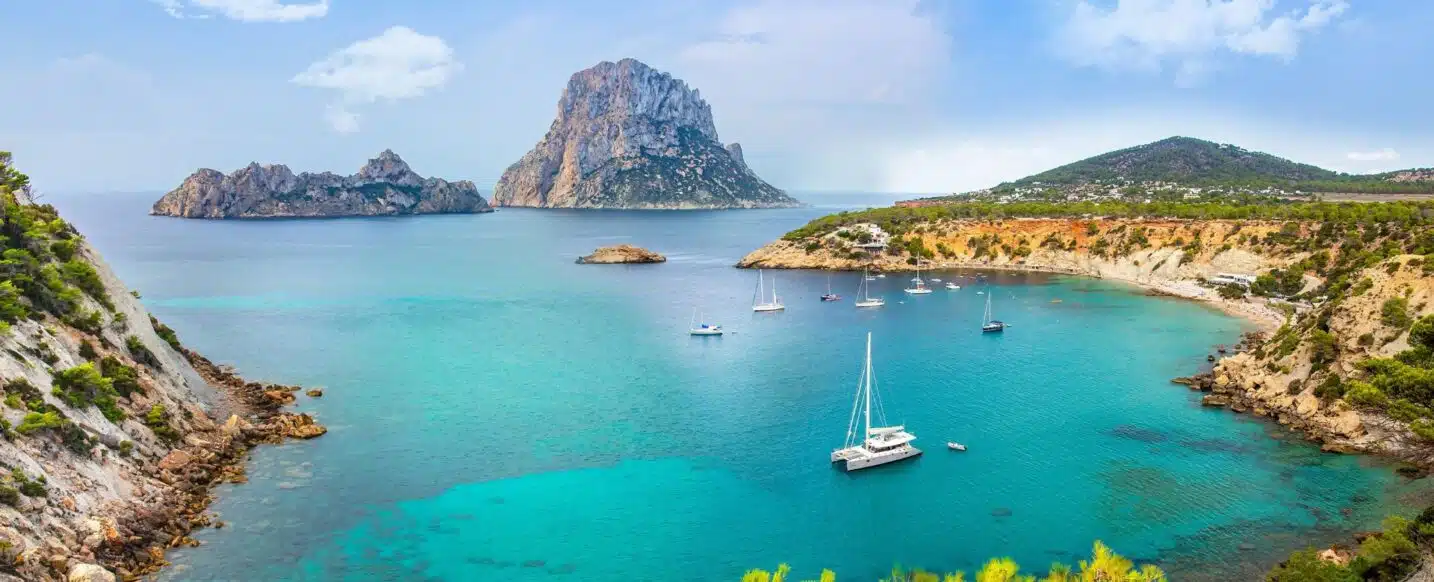 A panoramic view of a picturesque bay in Ibiza, Spain. The clear turquoise water is dotted with sailboats and yachts. The iconic Es Vedrà rock formation rises dramatically in the background against a backdrop of blue sky and fluffy white clouds.