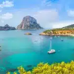 A panoramic view of a picturesque bay in Ibiza, Spain. The clear turquoise water is dotted with sailboats and yachts. The iconic Es Vedrà rock formation rises dramatically in the background against a backdrop of blue sky and fluffy white clouds.