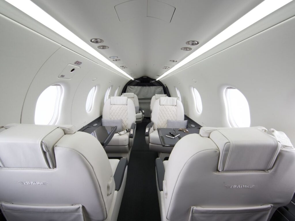 Interior view of a Pilatus PC-12 Turboprop aircraft featuring white leather seats, dark gray carpet, built-in side tables, and overhead lighting.