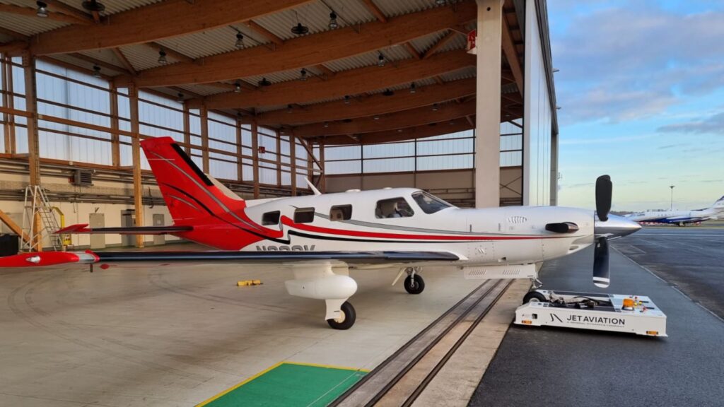 A white and red painted Piper PA-46-500TP Malibu Meridian turboprop aircraft with red, grey and black stripes over the side, parked inside a modern hangar with a wooden slat ceiling and a Jet Aviation pushback tug in the foreground.