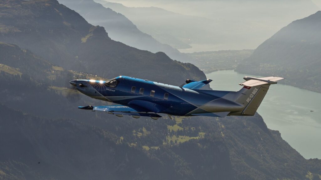 In-flight view of a blue and silver Pilatus PC-12 NGX single-engine turboprop, flying over a mountain lake with forested slopes and hazy peaks in the background.