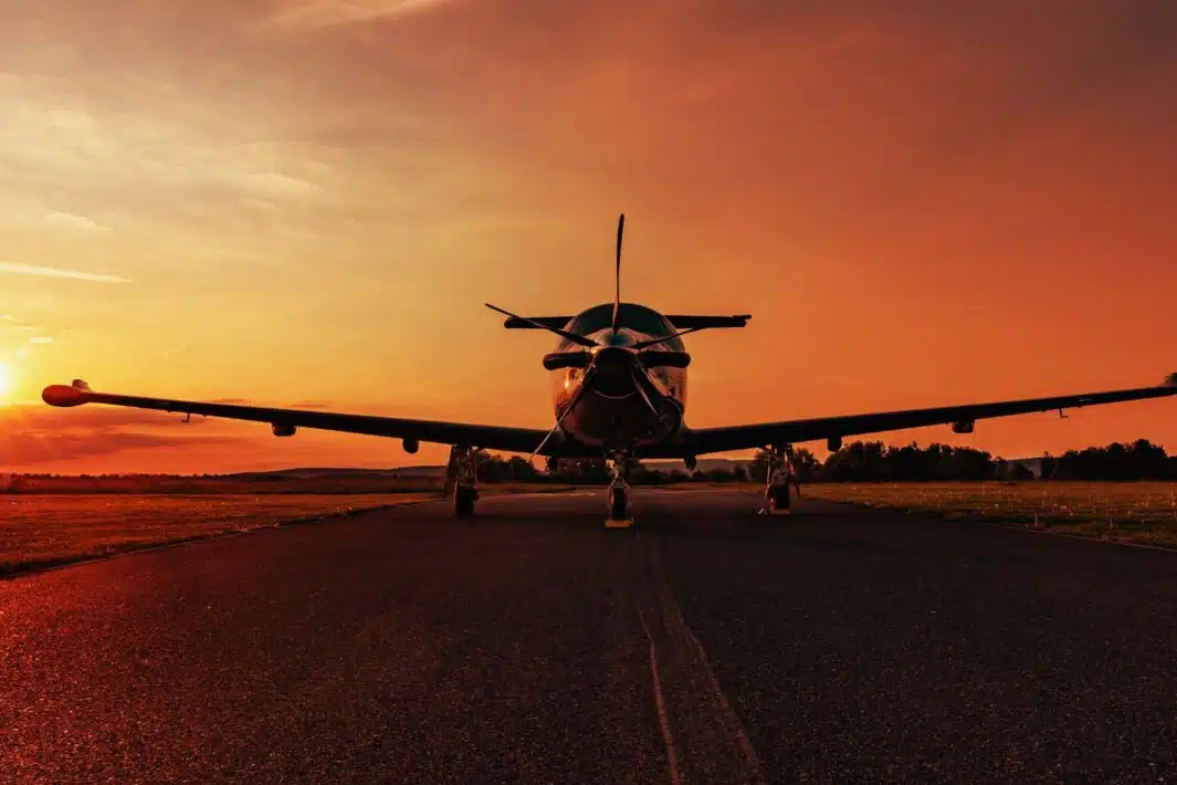 Front view of a Pilatus PC-12 at the airport, parked on an empty taxiway at sunset, with the sunburst directly behind the plane.