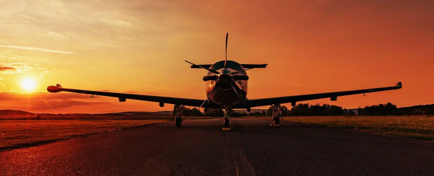 Front view of a Pilatus PC-12 at the airport, parked on an empty taxiway at sunset, with the sunburst directly behind the plane.