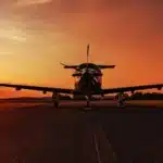 Front view of a Pilatus PC-12 at the airport, parked on an empty taxiway at sunset, with the sunburst directly behind the plane.