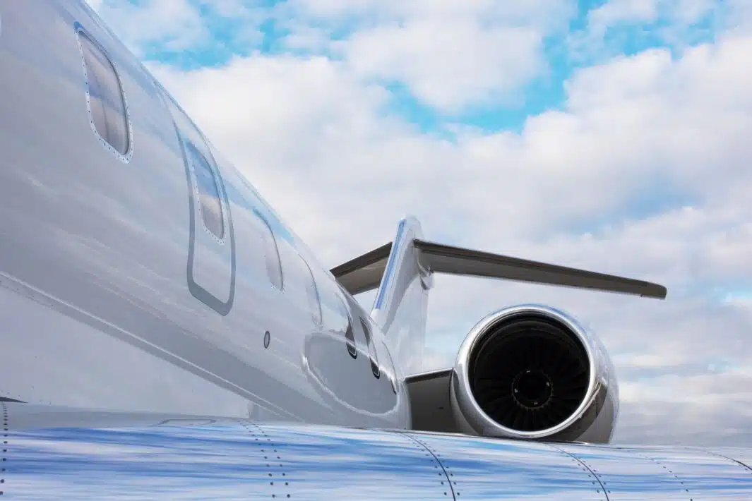 Close-up shot of a jet engine with visible turbine blades, attached to the side of a white airplane with windows. Blue sky and clouds are in the background.