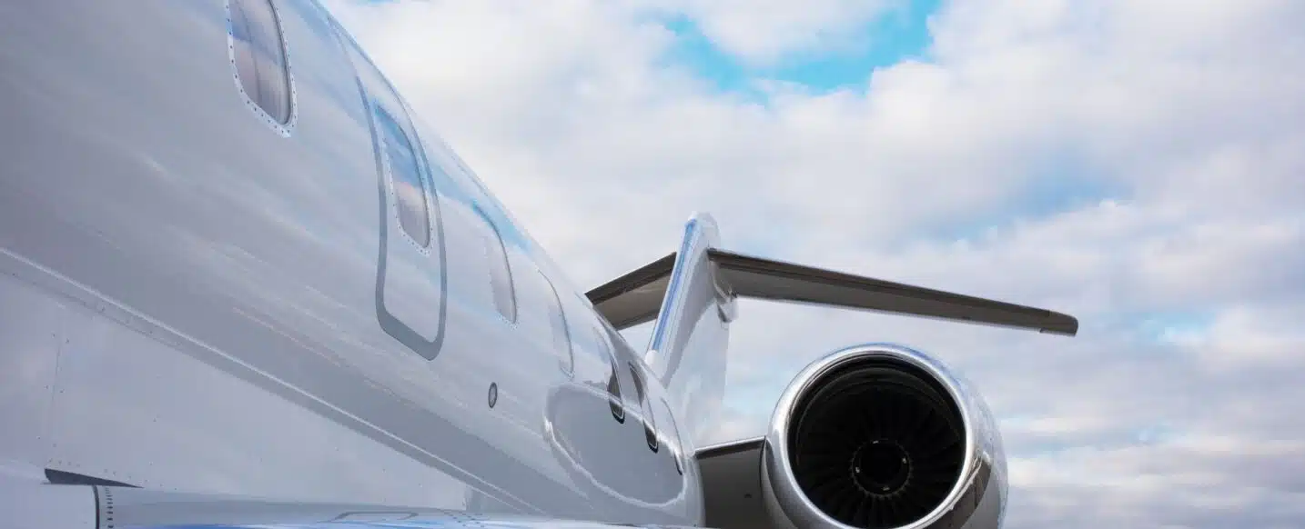 Close-up shot of a jet engine with visible turbine blades, attached to the side of a white airplane with windows. Blue sky and clouds are in the background.