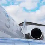 Close-up shot of a jet engine with visible turbine blades, attached to the side of a white airplane with windows. Blue sky and clouds are in the background.