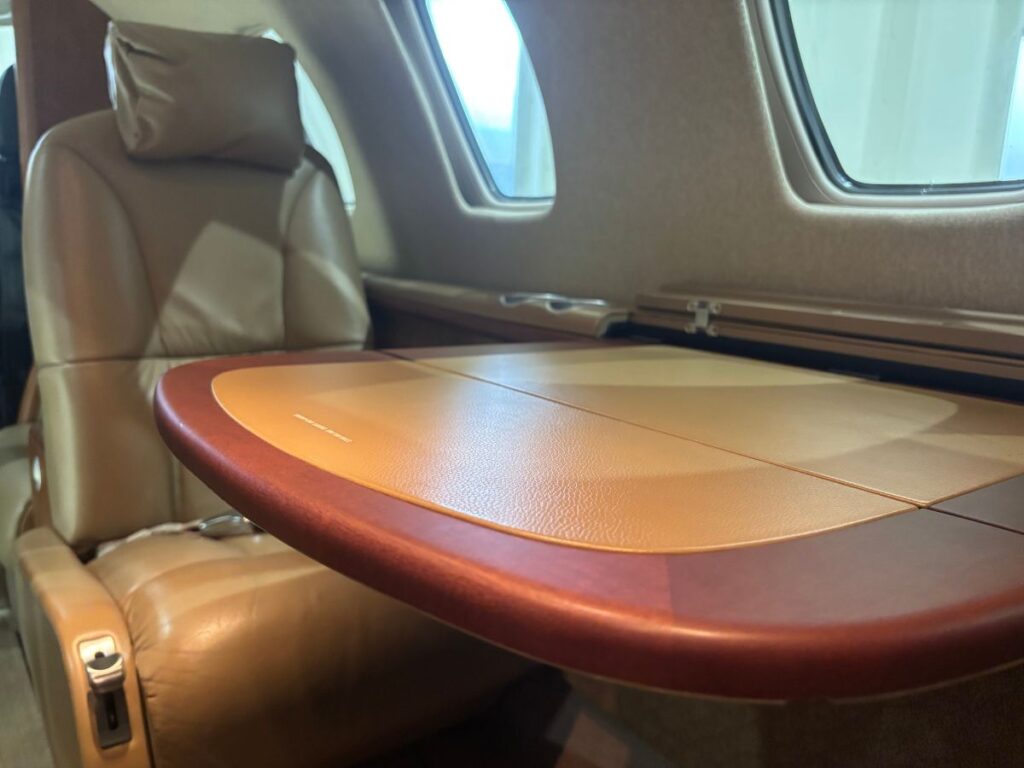 A close-up of the interior of a Cessna Citation CJ1+ private business jet, featuring a tan leather seat with a headrest, a partially extended folding table with a two-tone surface in light and dark tan, and a cabin window in the background. The seat shows a seatbelt and a side panel with cupholder.