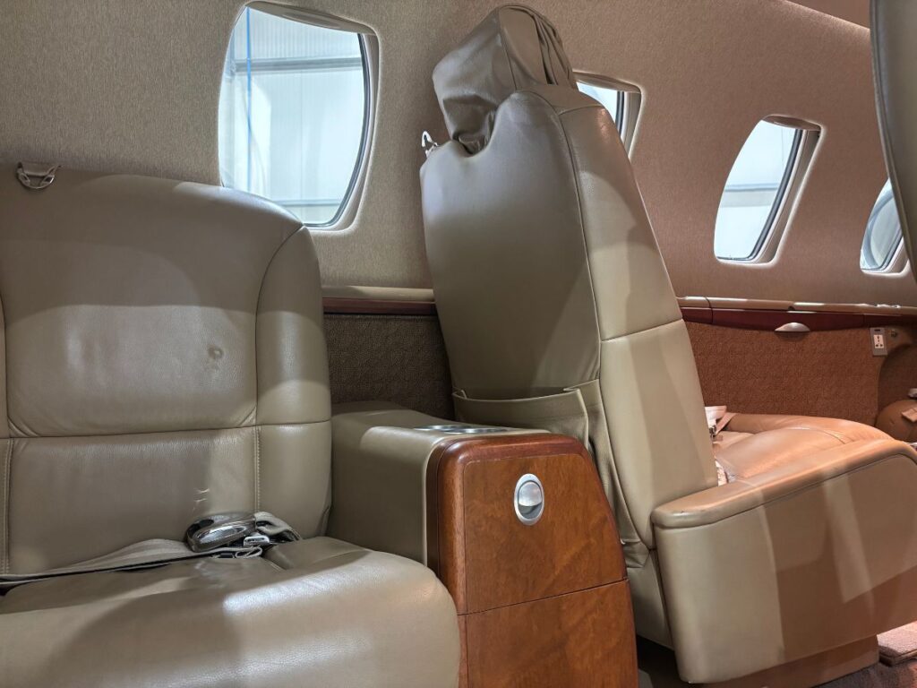 An image taken from the cabin door entrance of a CJ1+ private business jet, revealing the passenger seating area with two tan leather seats, a central armrest console with a wood finish and a silver button, and the characteristic oval-shaped windows. The seats are equipped with adjustable headrests, and the interior's design suggests a focus on comfort and functionality.