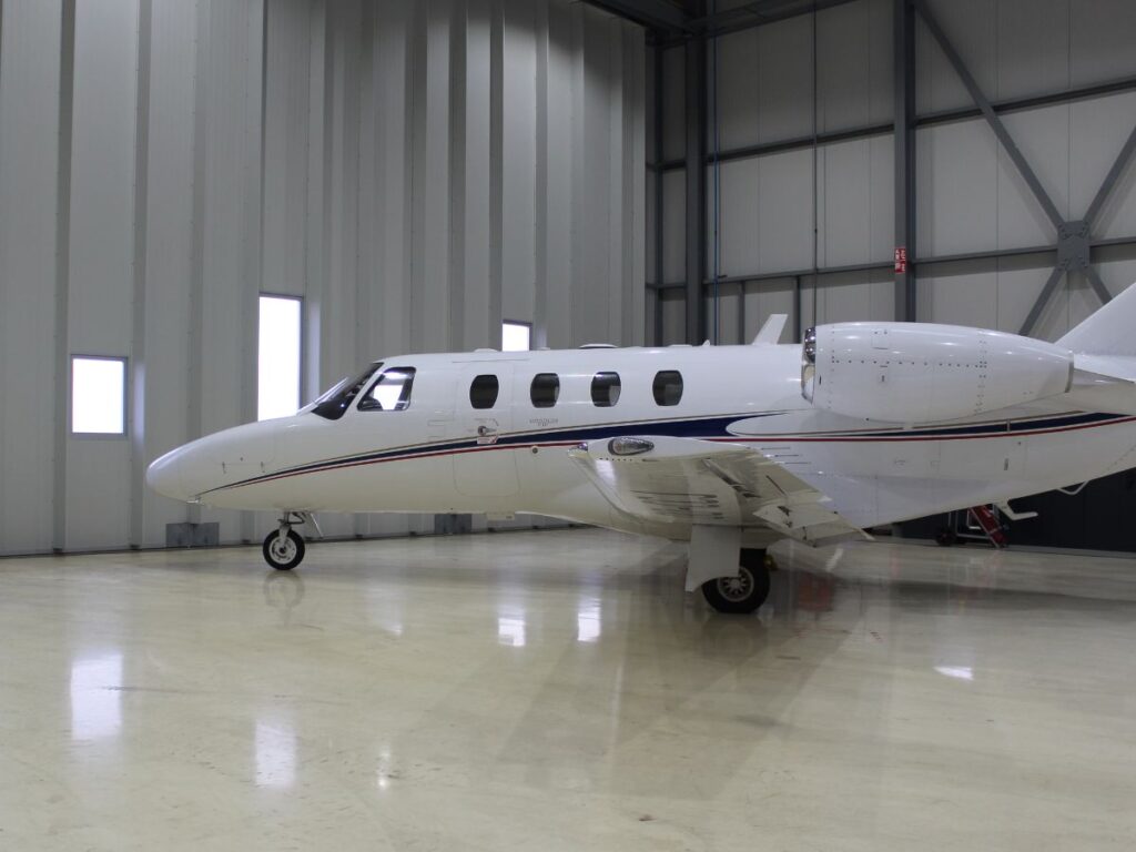 A white Cessna Citation CJ1+ corporate jet with a blue and red stripe parked on a shiny, light-colored floor inside a modern hangar. The hangar features white paneled walls, multiple rectangular windows, and visible structural beams.