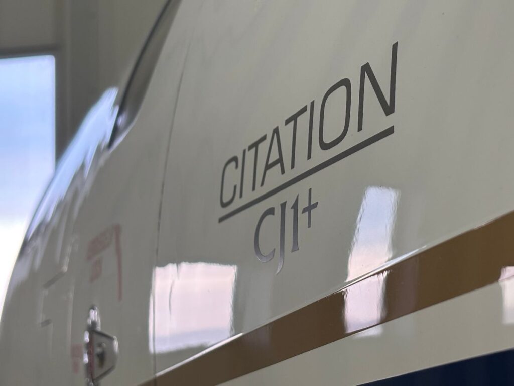 Close-up of the "CITATION CJ1+" lettering on the white fuselage of a Cessna Citation CJ1+ private business jet, showing the dark gray font and the reflective surface of the aircraft. The hangar door with windows in the background is slightly blurred.