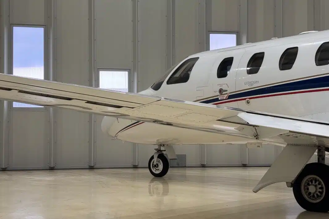 A white Cessna Citation CJ1+ private jet with a blue and red stripe along the fuselage, parked on a polished hangar floor. The image shows the left wing, landing gear, and a portion of the fuselage, including the "CITATION" lettering and several cabin windows. The hangar features white paneled walls and multiple rectangular windows.