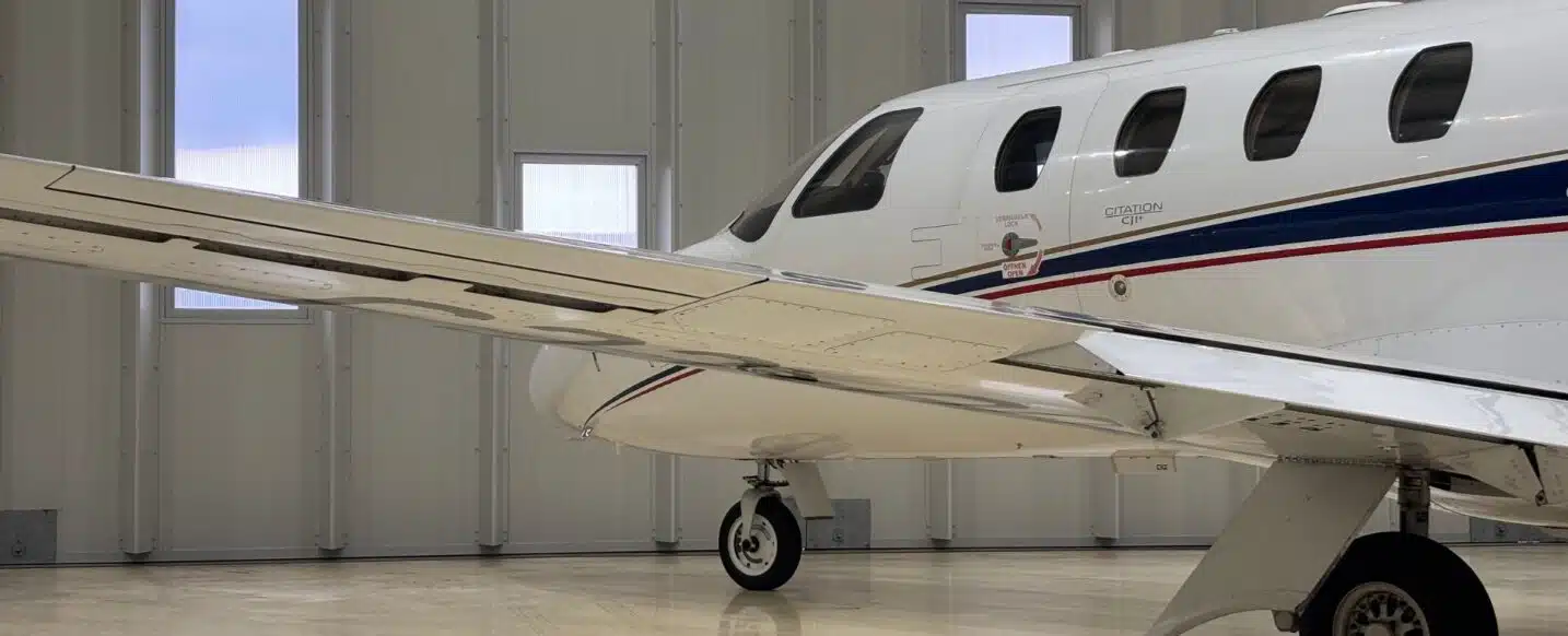 A white Cessna Citation CJ1+ private jet with a blue and red stripe along the fuselage, parked on a polished hangar floor. The image shows the left wing, landing gear, and a portion of the fuselage, including the "CITATION" lettering and several cabin windows. The hangar features white paneled walls and multiple rectangular windows.