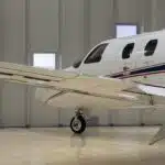 A white Cessna Citation CJ1+ private jet with a blue and red stripe along the fuselage, parked on a polished hangar floor. The image shows the left wing, landing gear, and a portion of the fuselage, including the "CITATION" lettering and several cabin windows. The hangar features white paneled walls and multiple rectangular windows.