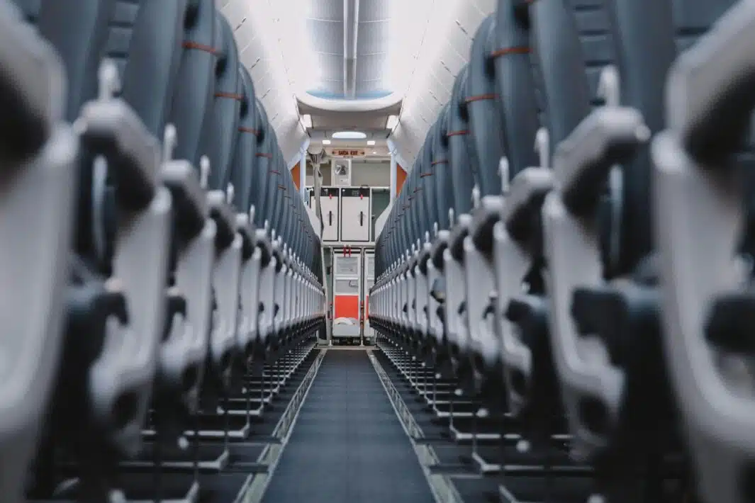 Empty airplane seats with dark gray upholstery and light gray headrests, lining a narrow aisle leading towards a service area.