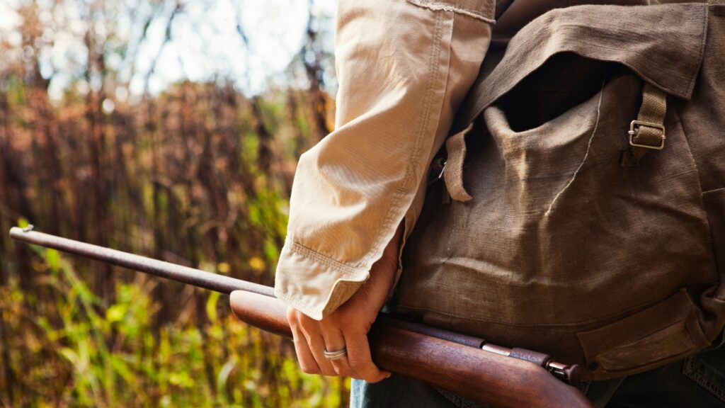 A close-up of a person's torso and hand holding a wooden rifle, angled downwards. The person wears a light beige jacket and a brown canvas backpack. The background is a blurred, natural setting with trees and foliage, suggesting an outdoor or hunting context. The focus is on the rifle and the person's grip.