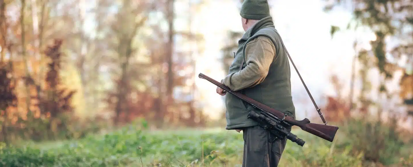 A man with a hunting weapon standing in grass and looking in to the forest