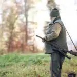 A man with a hunting weapon standing in grass and looking in to the forest