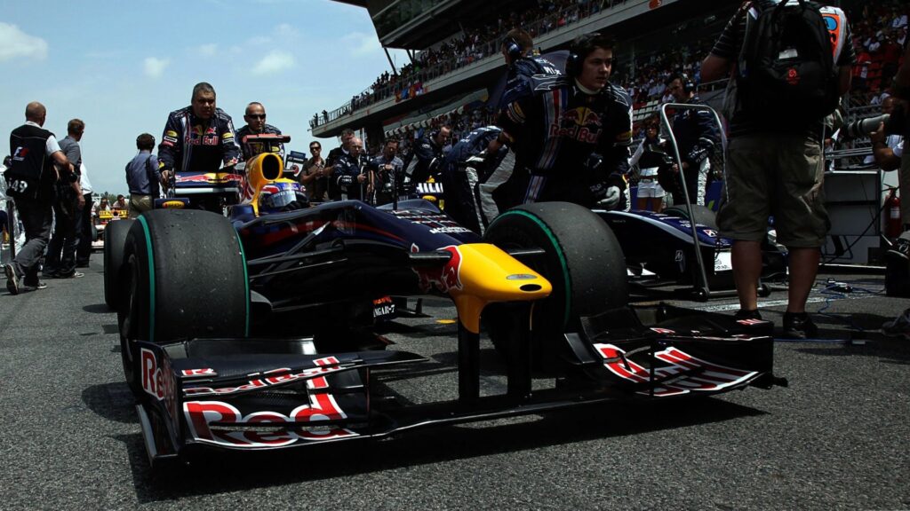 close up from low angle of a formula 1 racing car with red bull racing livery. the driver is sitting inside the racing car and the car is pushed by red bull racing team members. the tarmac is filled with people and in the background is a raised two-level platform tribune filled with people