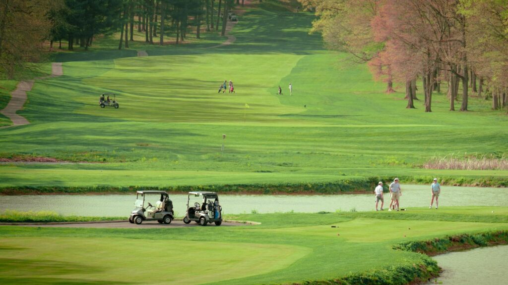 a serene and expansive golf course on a bright day. In the foreground, two golf carts are parked near a small body of water, with a few golfers preparing for a shot on the green. The lush, rolling fairways stretch into the distance, framed by tall trees with hints of spring foliage. Farther along the course, more golfers and carts can be seen, highlighting the leisurely and social nature of the game. The landscape is well-manicured, offering a peaceful, scenic atmosphere ideal for a day of golf.