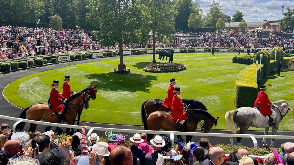 a vibrant scene at a prestigious  Royal Ascot horse racing event. The foreground shows riders in traditional red uniforms on horseback parading around a lush green paddock. The paddock is beautifully landscaped, featuring a central tree, manicured hedges, and a black horse statue surrounded by flowers. The grandstands are packed with spectators dressed in formal attire and elaborate hats, indicating a high-society occasion. The atmosphere is festive, elegant, and quintessentially British.