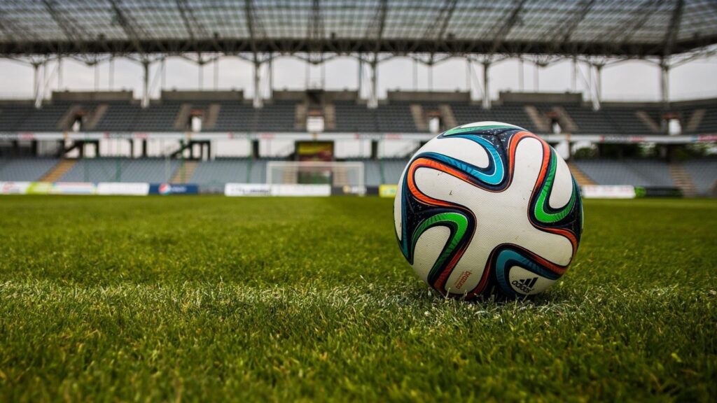 close-up view of a colorful Adidas soccer ball resting on a well-maintained grass field in a modern, empty stadium. The seating stands and a goalpost are visible in the background under a large, partially covered roof structure. The photo is taken from a low angle, emphasizing the ball in the foreground.