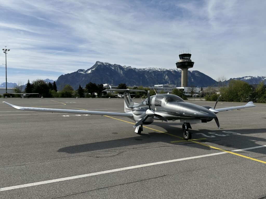 outside of a diamond da62 airplane parked at the airport with the air traffic control tower and mountainous terrain in the background.