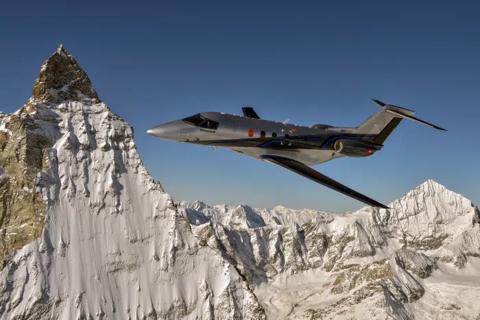 Pilatus PC-24 in grey livery with black stripes flying above mountains.