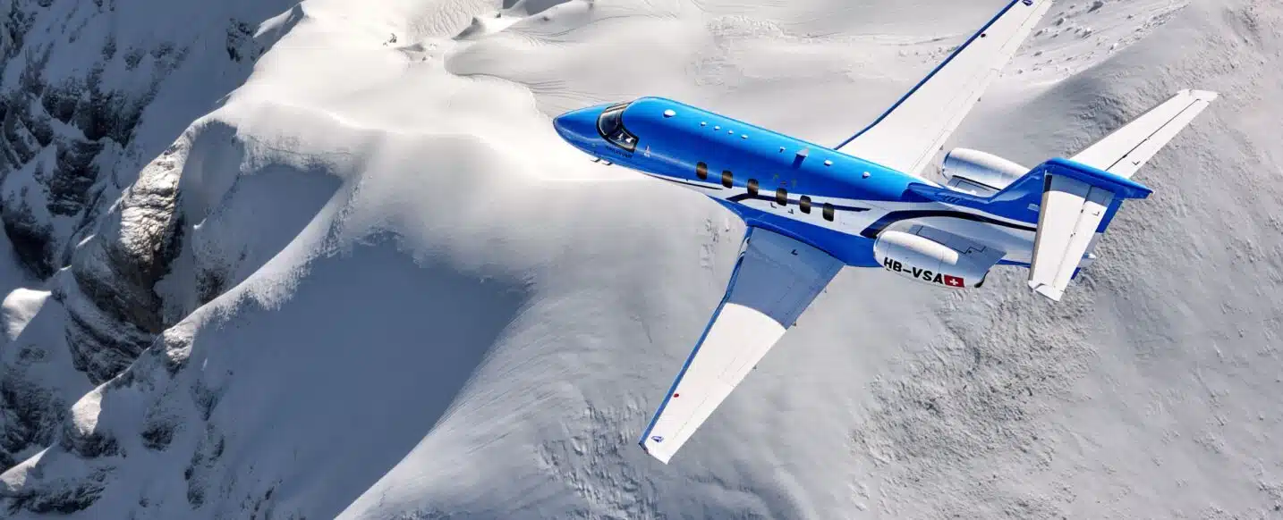 Top-down view of a blue Pilatus PC-24 flying above mountains.